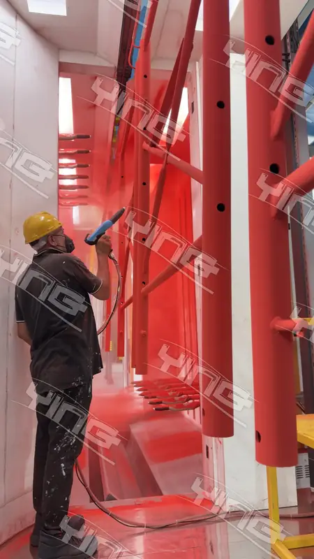 Operator spraying red powder coating by hand onto tall hanging parts inside a vertical booth